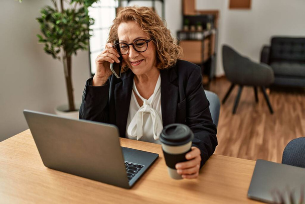 Mulher de negócios caucasiana de meia idade usando laptop e falando no smartphone no escritório. - Foto, Imagem