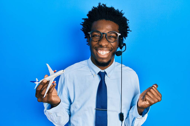 Genç Afro-Amerikan seyahat acentesi elinde uçağı tutan adam gururla bağırıyor, zaferi kutluyor ve kollarını kaldırdığı için çok heyecanlı.  - Fotoğraf, Görsel