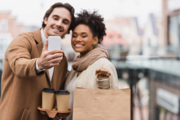 couple multiethnique flou avec des tasses en papier et sac à provisions prendre selfie dans le centre commercial  - Photo, image