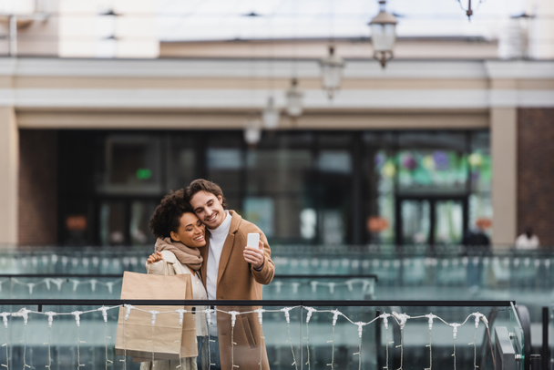 Kâğıt bardaklar ve alışveriş torbalarıyla alışveriş merkezinde selfie çeken neşeli çoklu etnik çift.  - Fotoğraf, Görsel