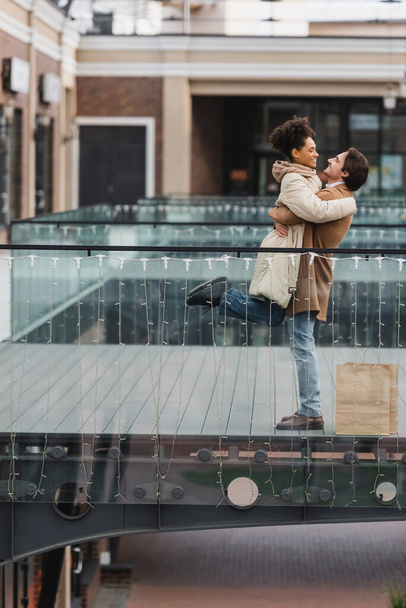 side view of happy man lifting african american girlfriend in shopping mall  - Fotoğraf, Görsel