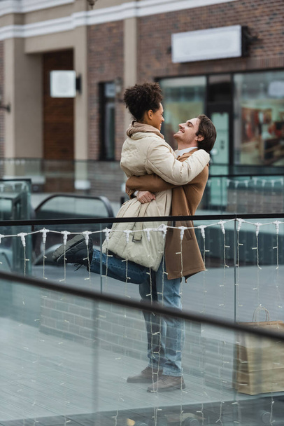cheerful man lifting african american girlfriend in shopping mall  - Photo, Image