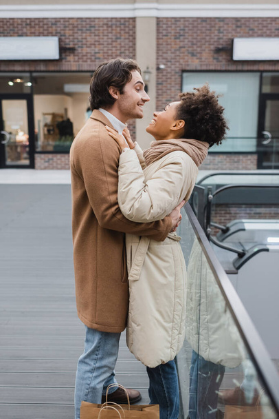 side view of happy man hugging cheerful african american girlfriend in shopping mall  - Photo, Image