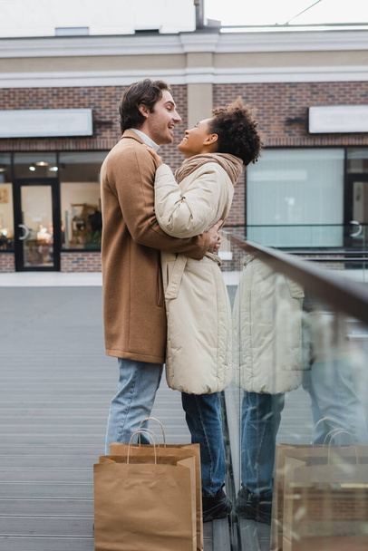 Seitenansicht von glücklichen Mann umarmt fröhliche afrikanisch-amerikanische Freundin in der Nähe von Einkaufstaschen in Mall  - Foto, Bild