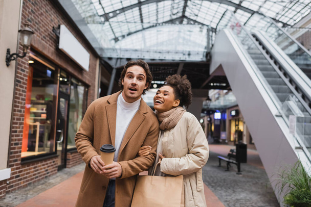 amazed multiethnic couple with purchases walking in mall  - Valokuva, kuva