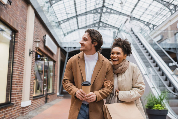 cheerful multiethnic couple with purchases walking in shopping mall  - Foto, Bild