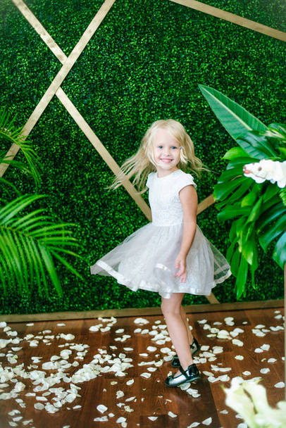  Little cute girl with blond hair in a white dress  and white flowers, lilies and orchids on a  background with a green tropical plants - Fotoğraf, Görsel