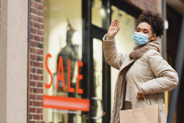 morena afro-americana em máscara médica segurando saco de compras e acenando a mão no shopping  - Foto, Imagem