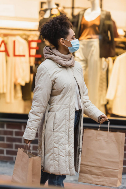 brunette african american woman in medical mask walking with shopping bags in mall  - Foto, afbeelding