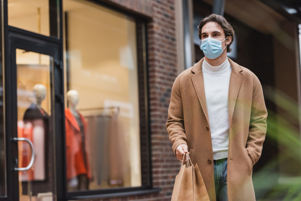 young man in medical mask and beige coat walking with purchases in shopping center - Zdjęcie, obraz