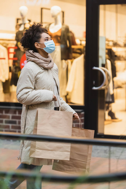 joven afroamericana mujer en máscara médica caminando con compras en el centro comercial - Foto, imagen
