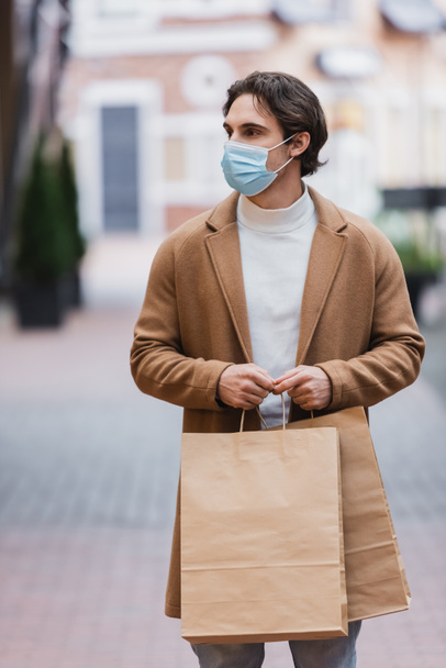 jeune homme en masque médical et manteau beige tenant achat dans le centre commercial - Photo, image