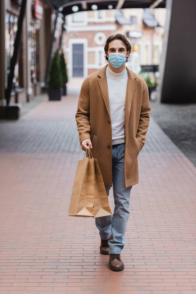 Medikal maskeli ve ceketli bir adam alışveriş merkezinde poşetlerle yürüyor. - Fotoğraf, Görsel