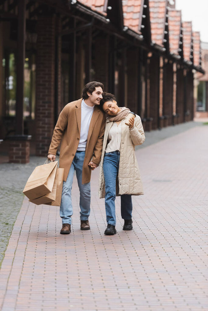aimant couple multiethnique en manteaux tenant la main tout en marchant avec des achats près du centre commercial - Photo, image