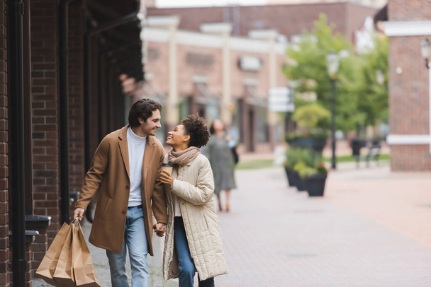 joyful multiethnic couple in coats holding hands while looking at each other and walking with purchases - Photo, Image