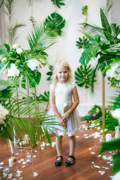  Little cute girl with blond hair in a white dress  and white flowers, lilies and orchids on a  background with a green tropical plants - Valokuva, kuva