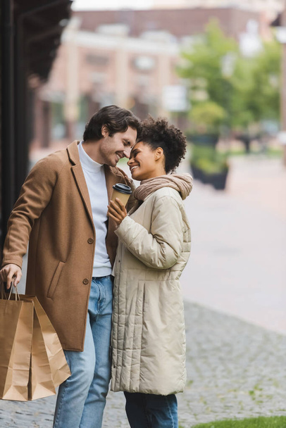pleased african american woman holding paper cup near smiling boyfriend with shopping bags - Foto, Imagem