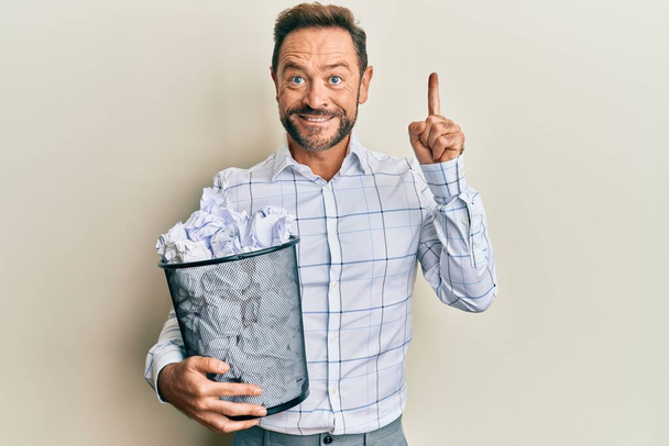 Мужчина средних лет держит в руках бумажную корзину со скомканными бумагами, улыбаясь идеей или вопросом, указывая пальцем со счастливым лицом, номер один  - Фото, изображение