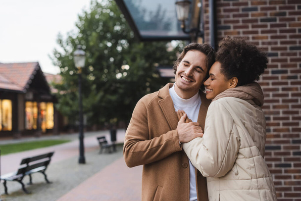 positive man embracing cheerful african american girlfriend near building outdoors  - Photo, Image