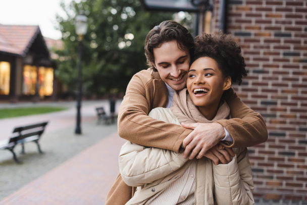 happy man embracing amazed african american girlfriend near building outdoors  - Photo, Image
