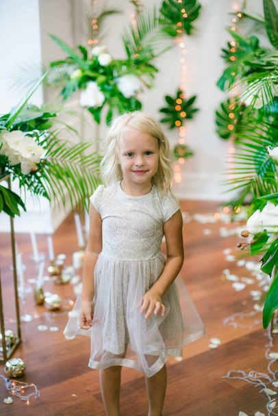  Menina bonito pouco com cabelo loiro em um vestido branco e flores brancas, lírios e orquídeas em um fundo com um verde plantas tropicais - Foto, Imagem