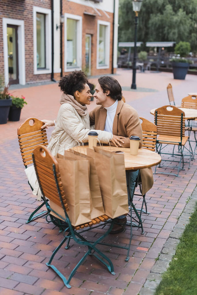 happy interracial couple sitting near paper cups and shopping bags in outdoor cafe  - Foto, imagen