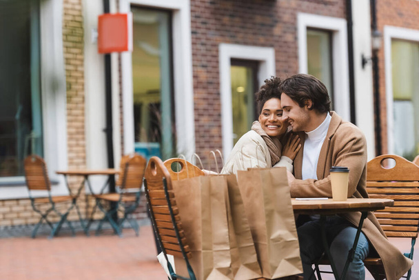 iloinen rotujenvälinen pari syleilee lähellä paperi cup ja ostoskassit ulkona kahvilassa  - Valokuva, kuva