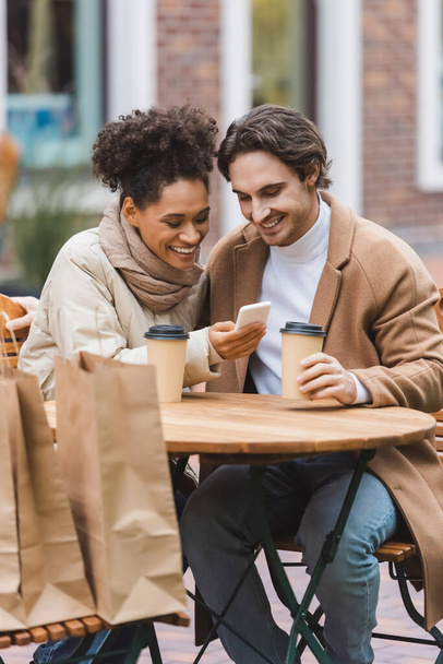 pleased african american woman holding smartphone near boyfriend with paper cup  - Foto, Bild