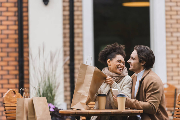 happy african american woman holding shopping bag and laughing near boyfriend with smartphone - Photo, Image