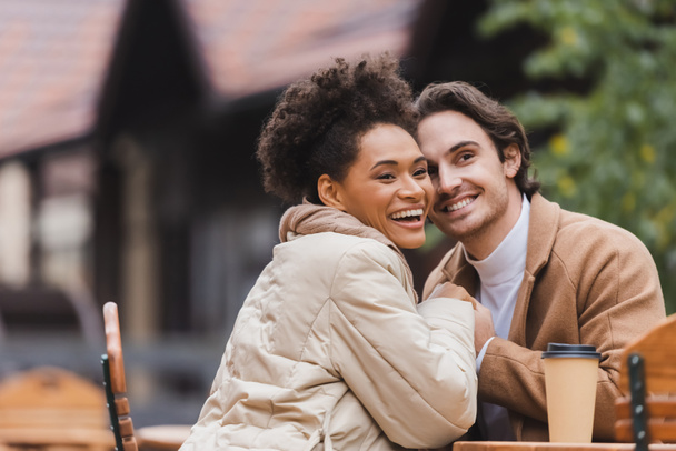 happy interracial couple in coats holding hands near paper cup on table - Fotó, kép