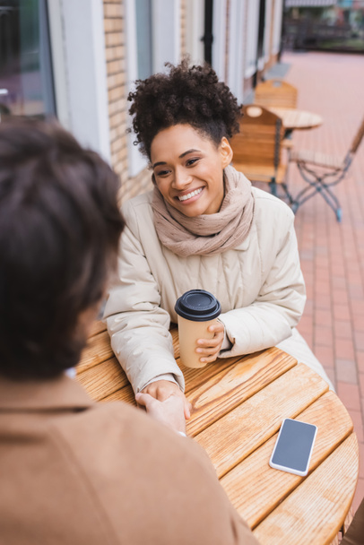 happy african american woman holding hands with blurred boyfriend near smartphone and paper cup  - Foto, Imagem