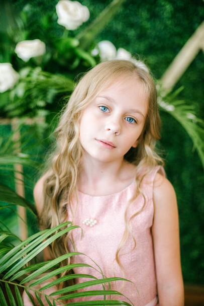  ragazza carina con lunghi capelli biondi in un abito rosa e fiori bianchi, gigli e orchidee su uno sfondo con piante tropicali verdi - Foto, immagini