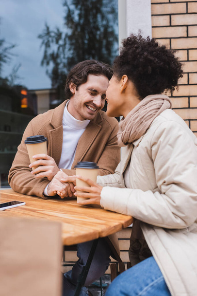 happy interracial couple holding hands near paper cups and blurred shopping bag - Photo, Image