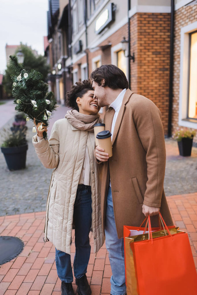 cheerful interracial couple with coffee to go, christmas shopping bags and small pine near mall  - 写真・画像