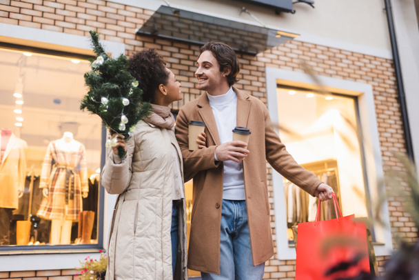 ευτυχισμένο πολυεθνικό ζευγάρι με καφέ για να πάει, τσάντες για ψώνια Χριστουγέννων και μικρό έλατο κοντά στο εμπορικό κέντρο  - Φωτογραφία, εικόνα