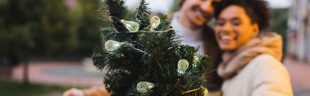 pareja interracial borrosa cerca de ramas de pino de Navidad con luces de guirnalda, pancarta - Foto, Imagen