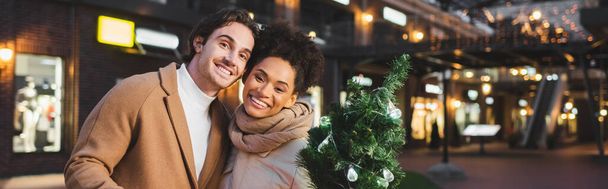 happy multiethnic couple holding small christmas pine in evening, banner - Photo, Image