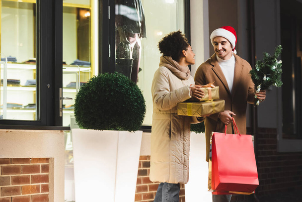 happy man in santa hat holding shopping bags and pine near african american girlfriend with gift boxes near mall - 写真・画像