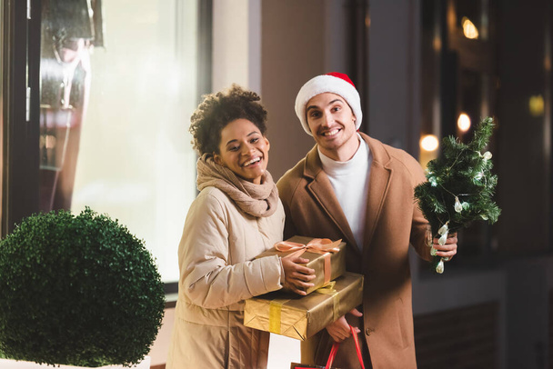 glücklicher Mann in Weihnachtsmannmütze mit Einkaufstaschen und kleiner Tanne in der Nähe einer afrikanisch-amerikanischen Freundin mit Geschenkschachteln  - Foto, Bild