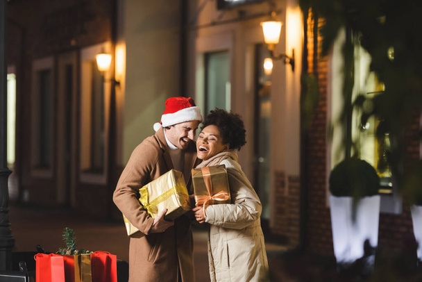 サンタ・ハットのハッピー・マンとアフリカ系アメリカ人のガールフレンドがクリスマスプレゼント箱を持ちながら笑っています - 写真・画像