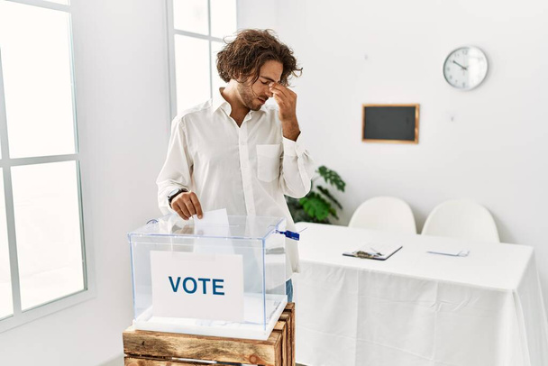 Молодий іспаномовний чоловік, який голосує, кладе конверт у коробку для голосування, втомився, потираючи ніс і очі, відчуваючи втому і головний біль. концепція стресу та розчарування
.  - Фото, зображення