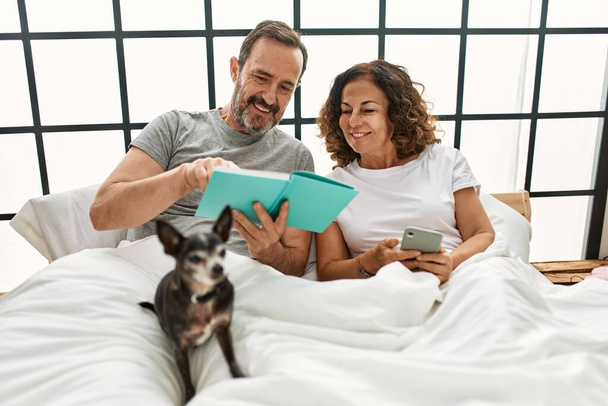 Середньовіччя його іспанська пара читає книгу і використовує смартфон. Лежачи на ліжку з собакою вдома
. - Фото, зображення