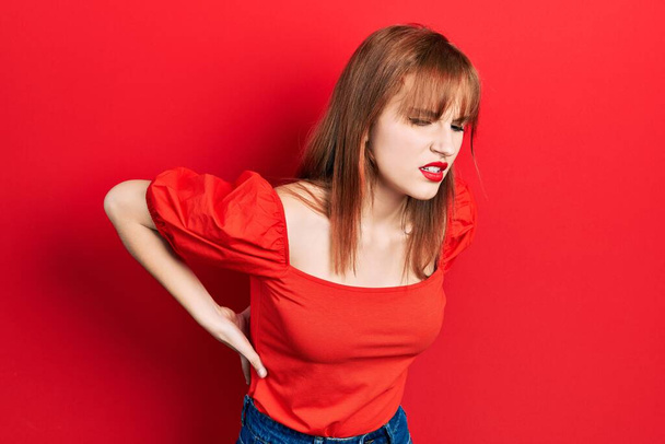 Рыжая молодая женщина в обычной красной футболке страдает от боли в спине, касаясь спины рукой, мышечной боли  - Фото, изображение