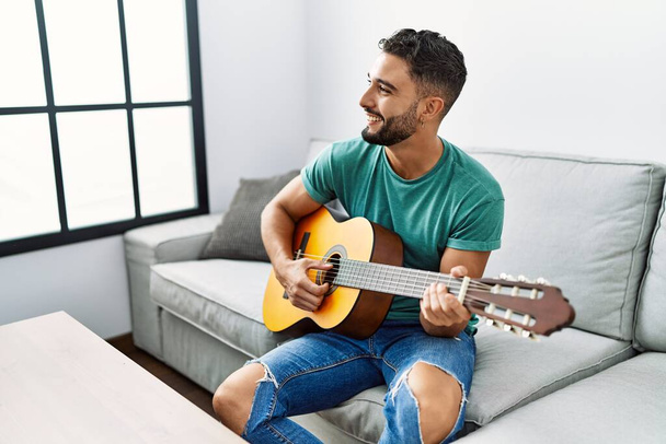 Young arab man playing classical guitar sitting on sofa at home - Foto, Imagem