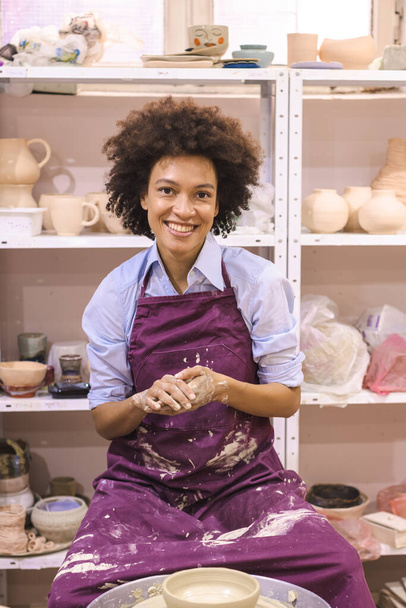 陶器の車輪の上に粘土を成形する創造的なアフロアメリカの若い女性。陶芸工房でのワークショップ. - 写真・画像