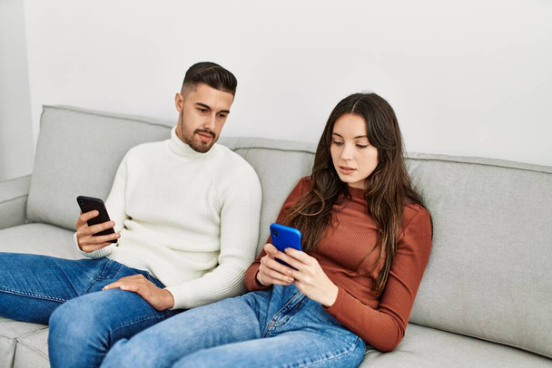 Молода іспаномовна пара використовує смартфон, що сидить на дивані вдома
. - Фото, зображення