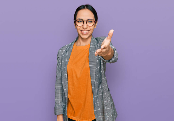 Молодая латиноамериканка в деловой куртке и очках, улыбаясь дружелюбно предлагая рукопожатие в качестве приветствия и приветствия. успешный бизнес.  - Фото, изображение