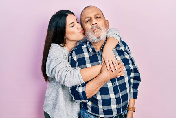 Young brunette woman and senior man standing over pink background. Daughter and father hugging and bonding together as happy family - Photo, Image