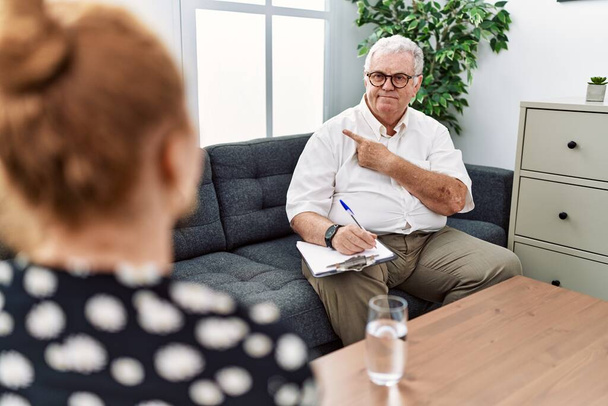 Hombre psicólogo sénior en la consulta señalando con el dedo de la mano a un lado mostrando publicidad, cara seria y tranquila  - Foto, imagen