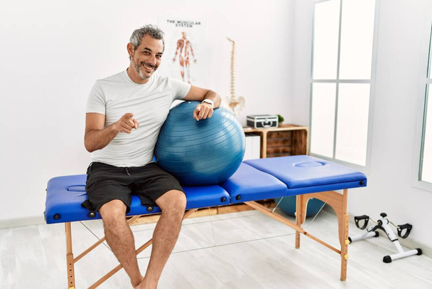 Hombre hispano de mediana edad en la clínica de recuperación del dolor sosteniendo la pelota de pilates señalándote a ti y a la cámara con los dedos, sonriendo positiva y alegre  - Foto, Imagen